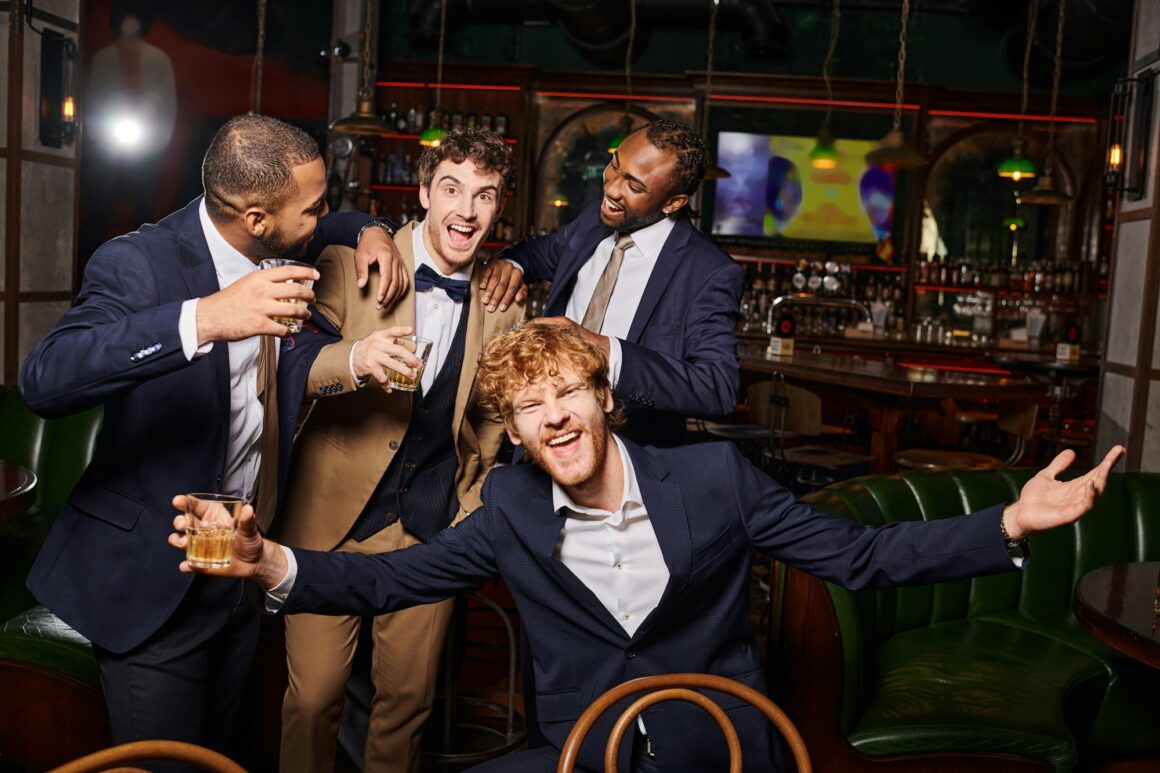 happy interracial friends in formal wear having bachelor party and holding whiskey in bar, diversity