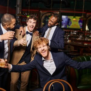 happy interracial friends in formal wear having bachelor party and holding whiskey in bar, diversity