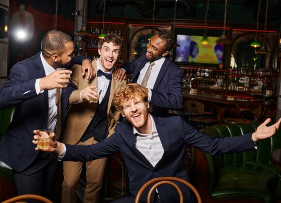 happy interracial friends in formal wear having bachelor party and holding whiskey in bar, diversity